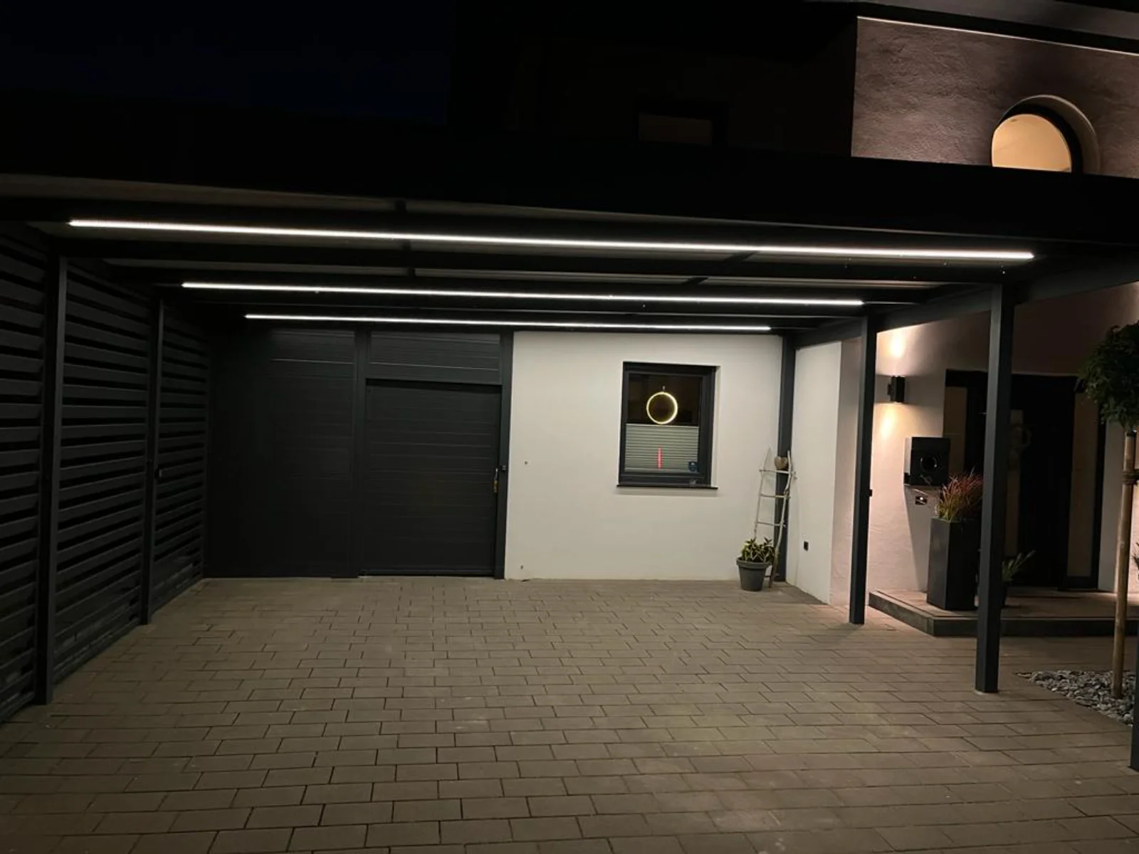 Überdachter Stellplatz mit modernem Metall-Carport und integrierten LED-Lichtschienen auf gepflasterter Fläche vor Hausfassade