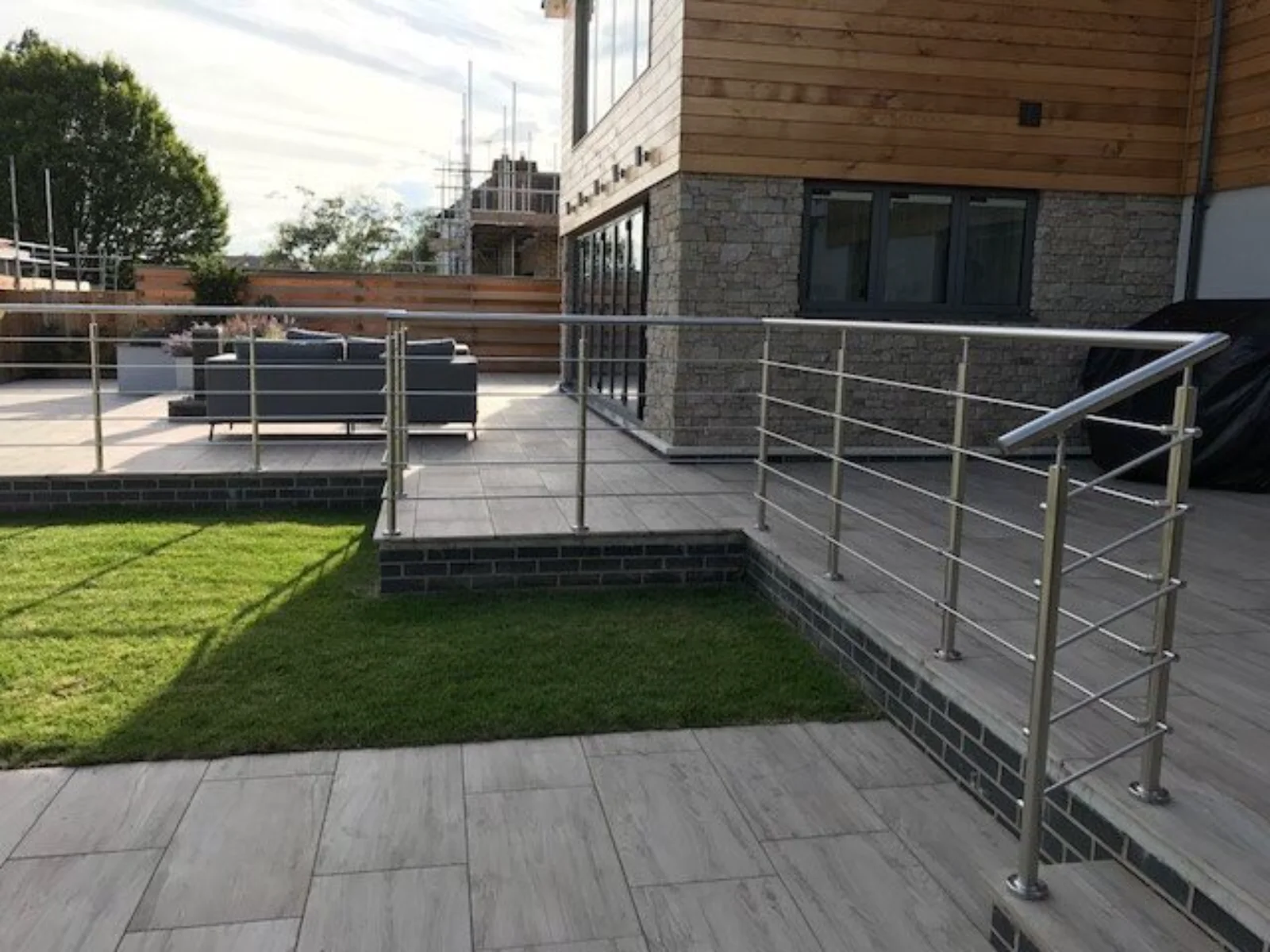 Terrassenpodest mit modernem Edelstahl-Metallgeländer, graue Bodenplatten und Rasen vor einer Holz-/Steinfassade