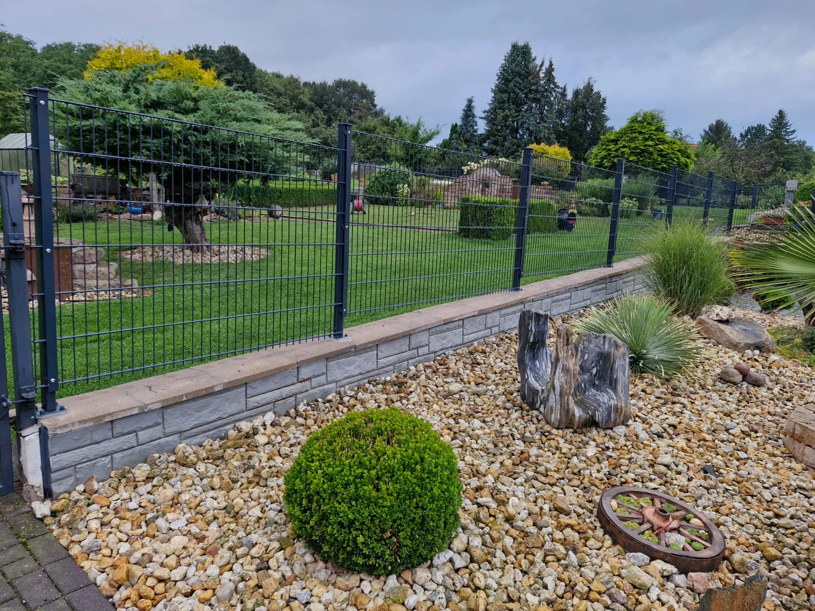 Moderner Metallzaun als Metallgeländer auf Natursteinmauer mit Kiesbeet und Bepflanzung im Garten