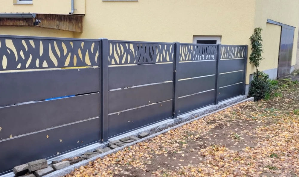Grauer Metallzaun mit dekorativen Ausschnitten vor gelber Hausfassade und Laub