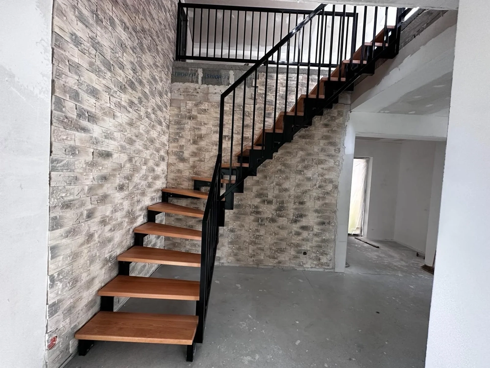 Freitragende Treppe aus Holz mit schwarzem Metallgeländer vor roher Ziegel- und Weißputzwand im Innenbereich