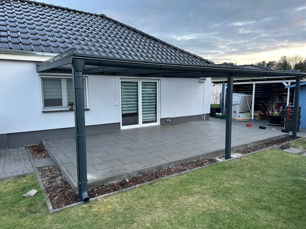 Anthrazitfarbene Terrassenüberdachung aus Aluminium mit integrierter Regenrinne über gepflasterter Terrasse vor weißer Hausfassade