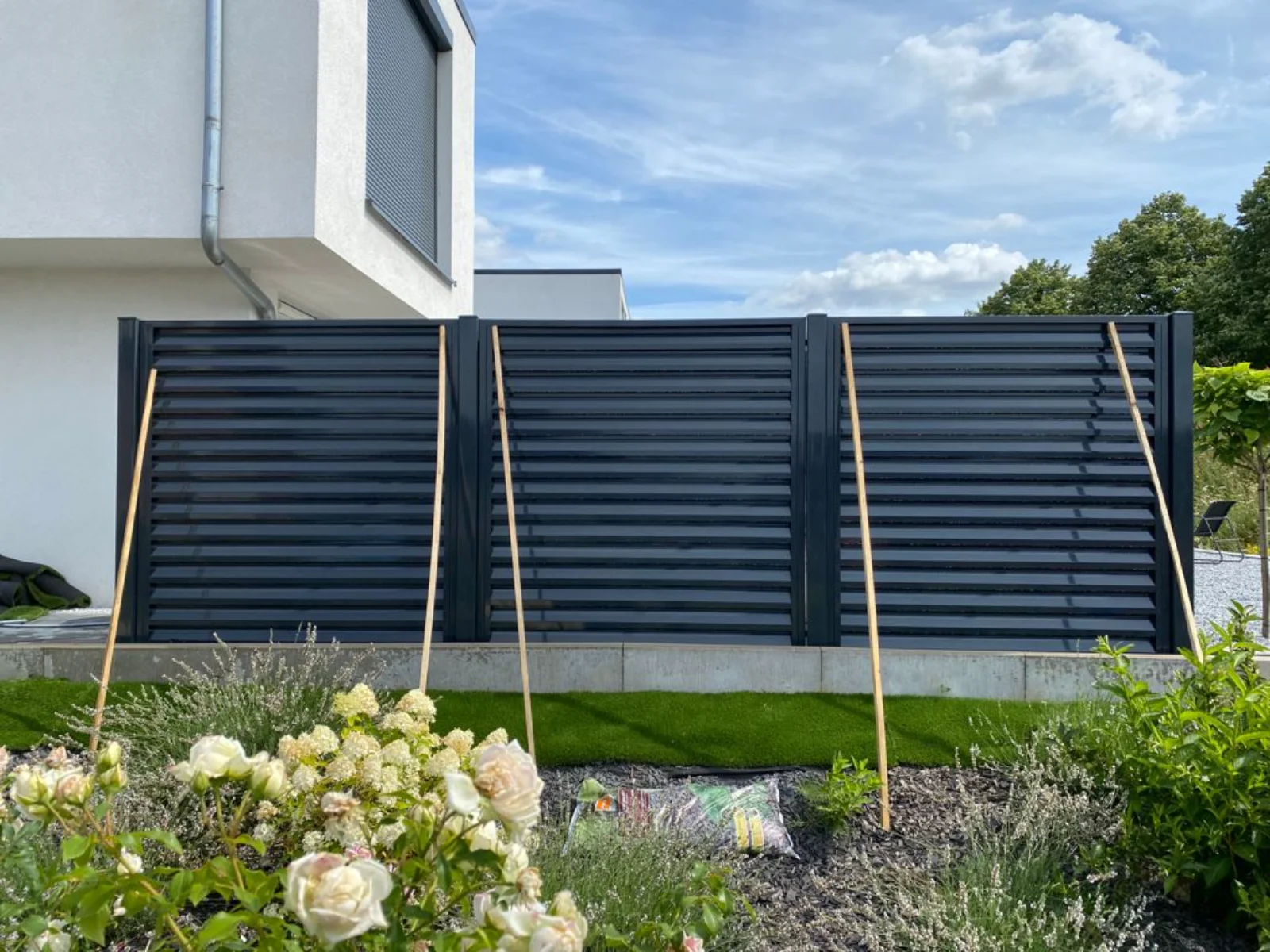 Drei horizontale Metallzaunelemente mit Lamellen vor modernem Haus, gestützt auf Stäbe