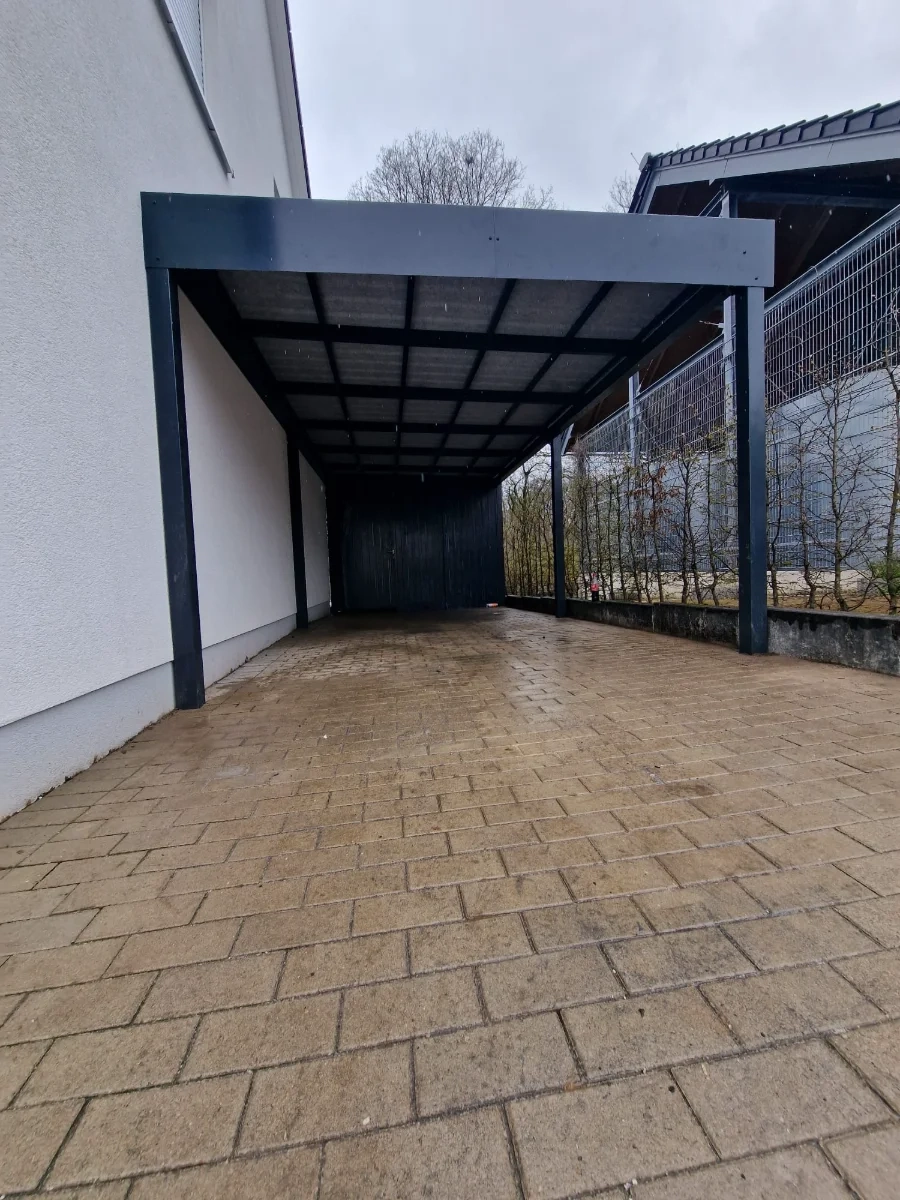 Moderner Carport aus schwarzem Stahl mit Pultdach und gepflasterter Zufahrt an Hauswand