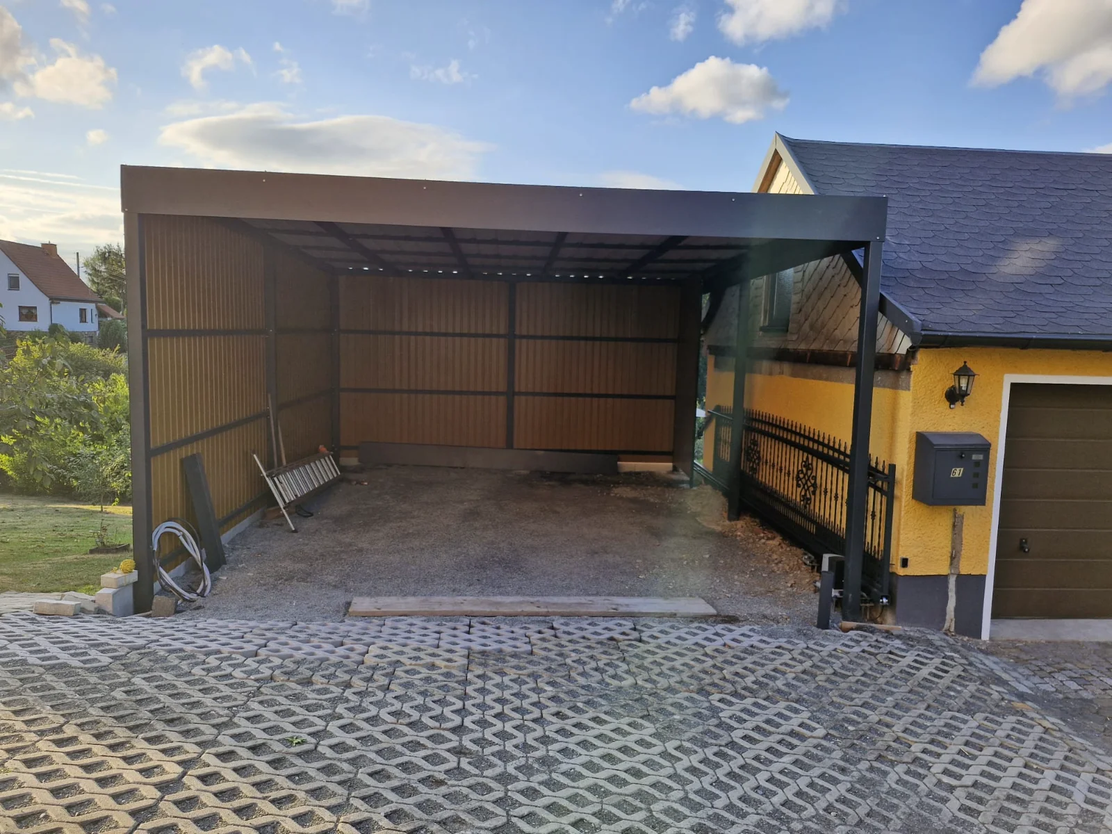 Überdachter Carport mit schwarzem Metallrahmen und Holzverkleidung neben gelbem Haus mit gepflasterter Auffahrt
