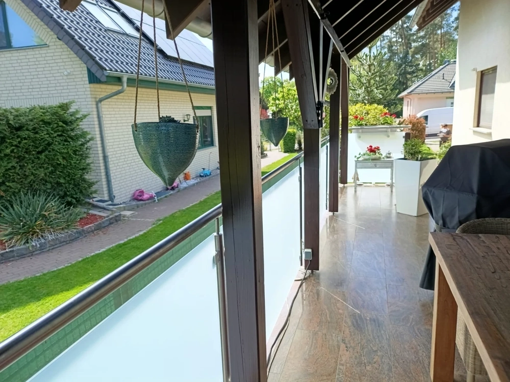 Terrassengeländer mit Edelstahlhandlauf, Milchglasfüllung und Holzkonstruktion auf einer sonnigen Terrasse mit Hängetöpfen