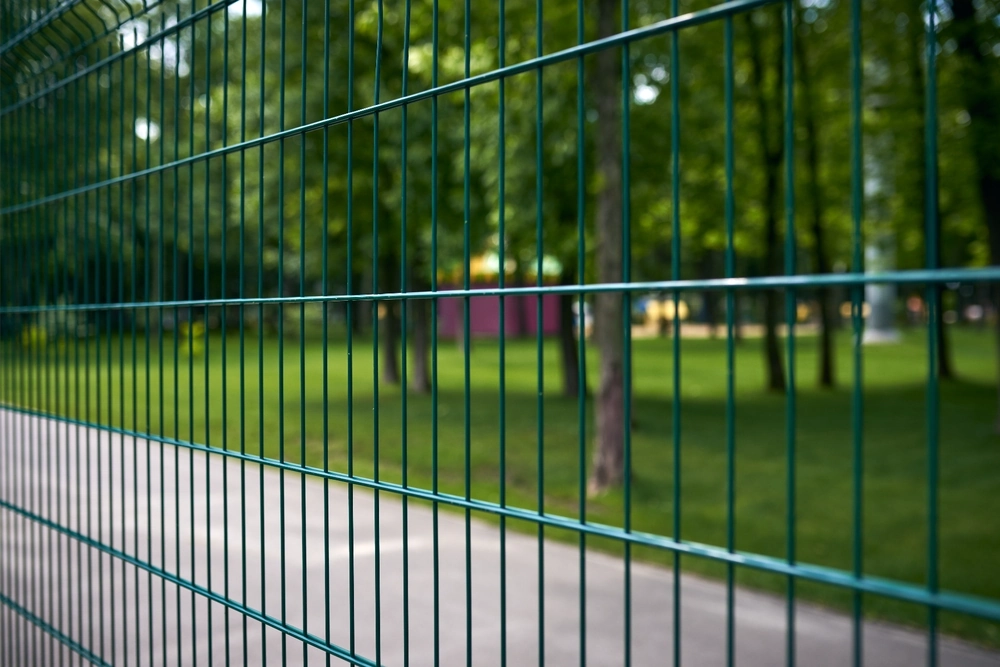 Grüner Doppelstabmattenzaun vor Parkweg und Bäumen im Hintergrund