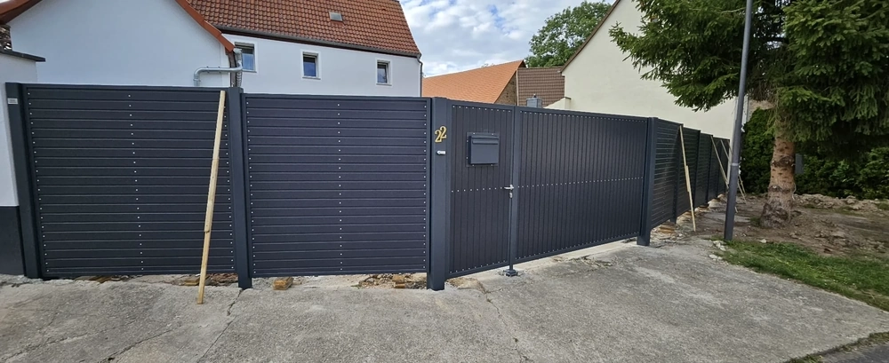 Dunkelgrauer Lamellenzaun mit integriertem Zugangstor auf Betonauffahrt vor Einfamilienhaus
