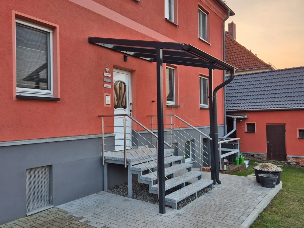 Terrassenüberdachung am Hauseingang mit Metallpfosten, Glasdach, Edelstahlgeländer und Granittreppe auf gepflastertem Weg