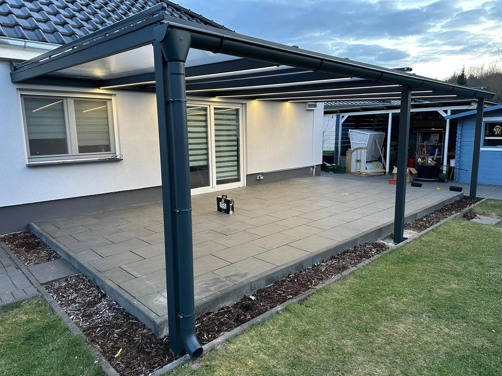 Moderne Terrassenüberdachung aus dunkelgrauem Metall mit LED-Beleuchtung über gefliester Terrasse vor Haus