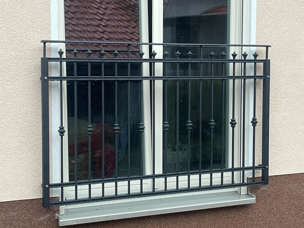 Schwarzes schmiedeeisernes Balkongeländer vor zweiflügeligem Fenster an heller Putzfassade