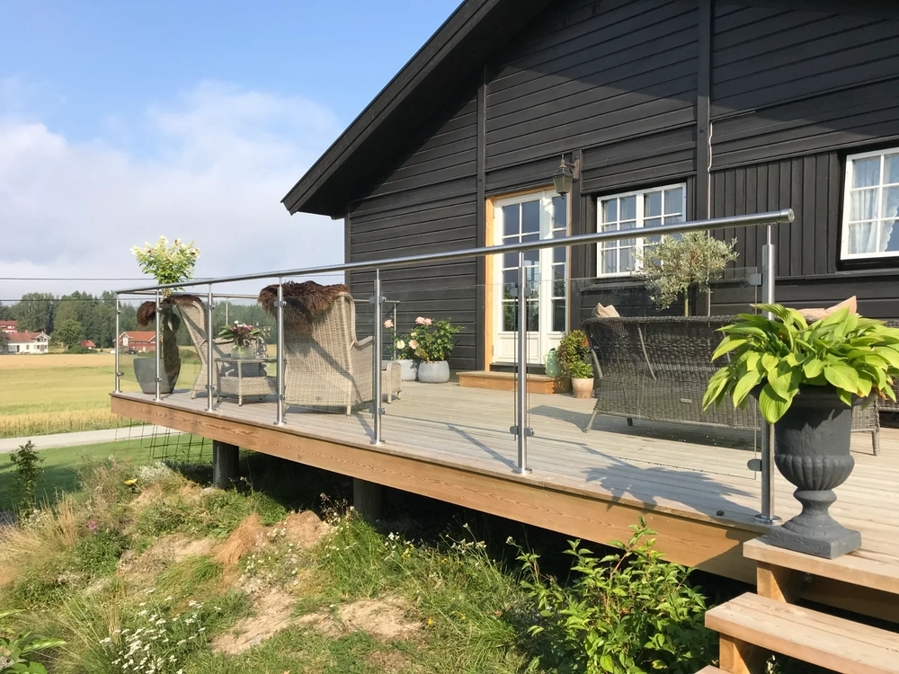 Terrassengeländer aus Edelstahl und Glas an moderner Holzterrasse mit Sitzmöbeln und Pflanzen vor schwarzem Haus